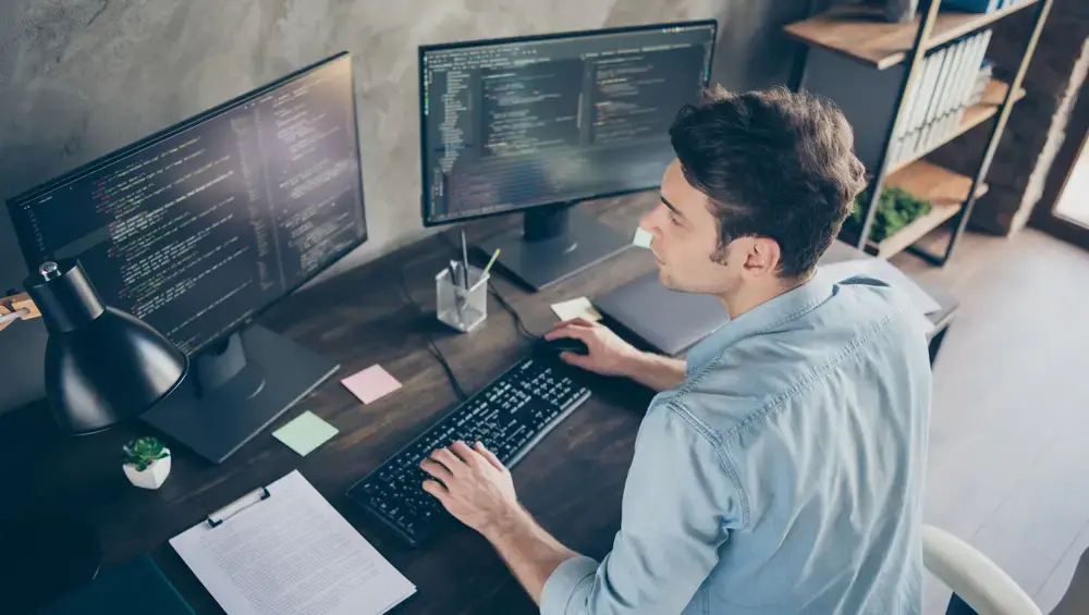man in front of two computer screens showing code
